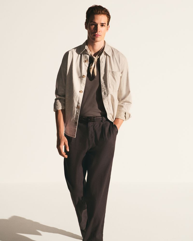 A young man in casual fashion stands in a photo studio wearing a tee shirt, jacket, trousers, and kerchief against a gradient white background.