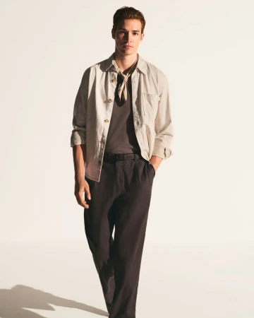 A young man in casual fashion stands in a photo studio wearing a tee shirt, jacket, trousers, and kerchief against a gradient white background.