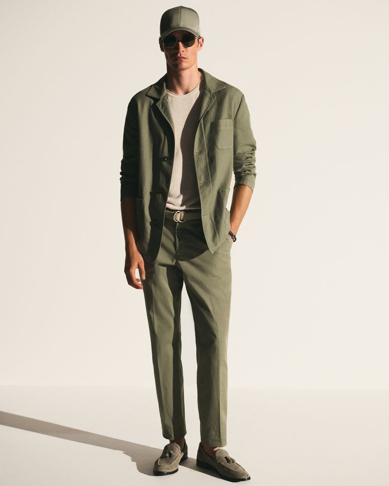 A full shot of a single adult male standing in a photo studio wearing sunglasses, a matching set, and tassel loafers.