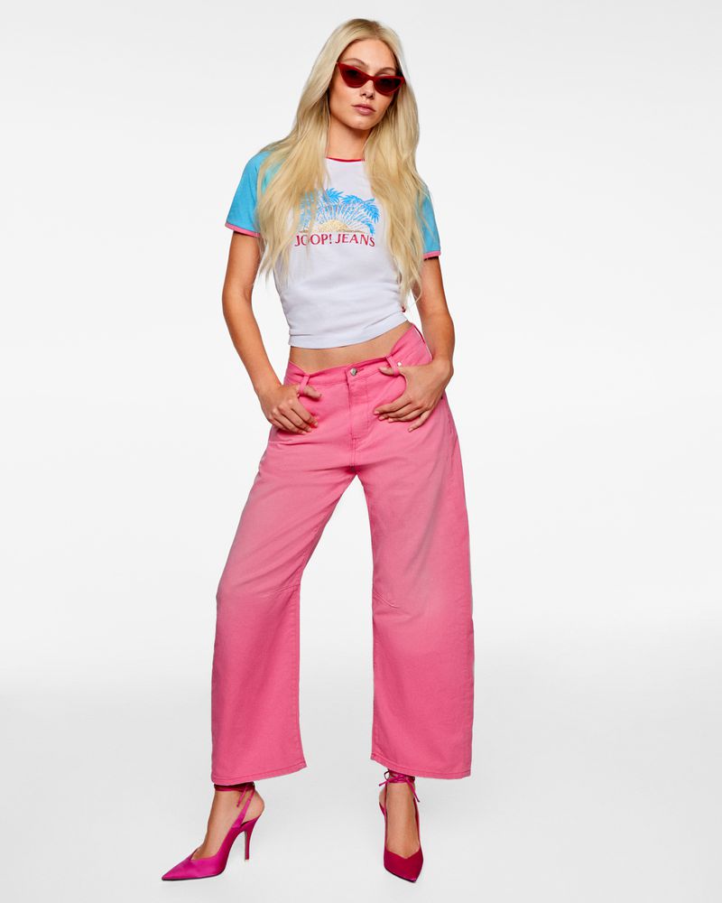 A woman with long blonde hair poses in bright pink jeans, a white t-shirt with a blue and red neckline, and red sunglasses on a white background.