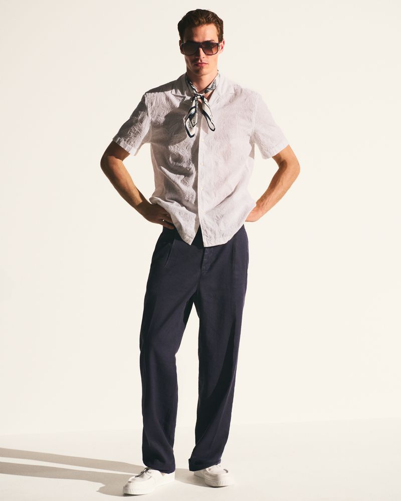 Full-length studio shot of a young adult male wearing a white short-sleeved shirt, navy blue pants, white sneakers, and sunglasses.