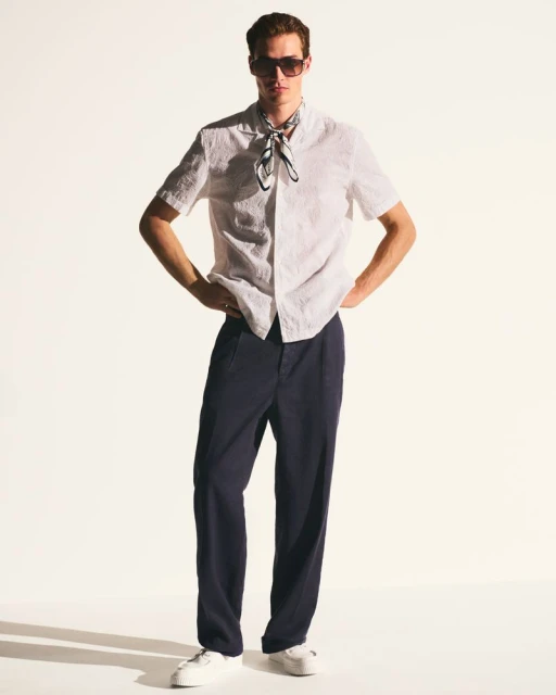 Full-length studio shot of a young adult male wearing a white short-sleeved shirt, navy blue pants, white sneakers, and sunglasses.