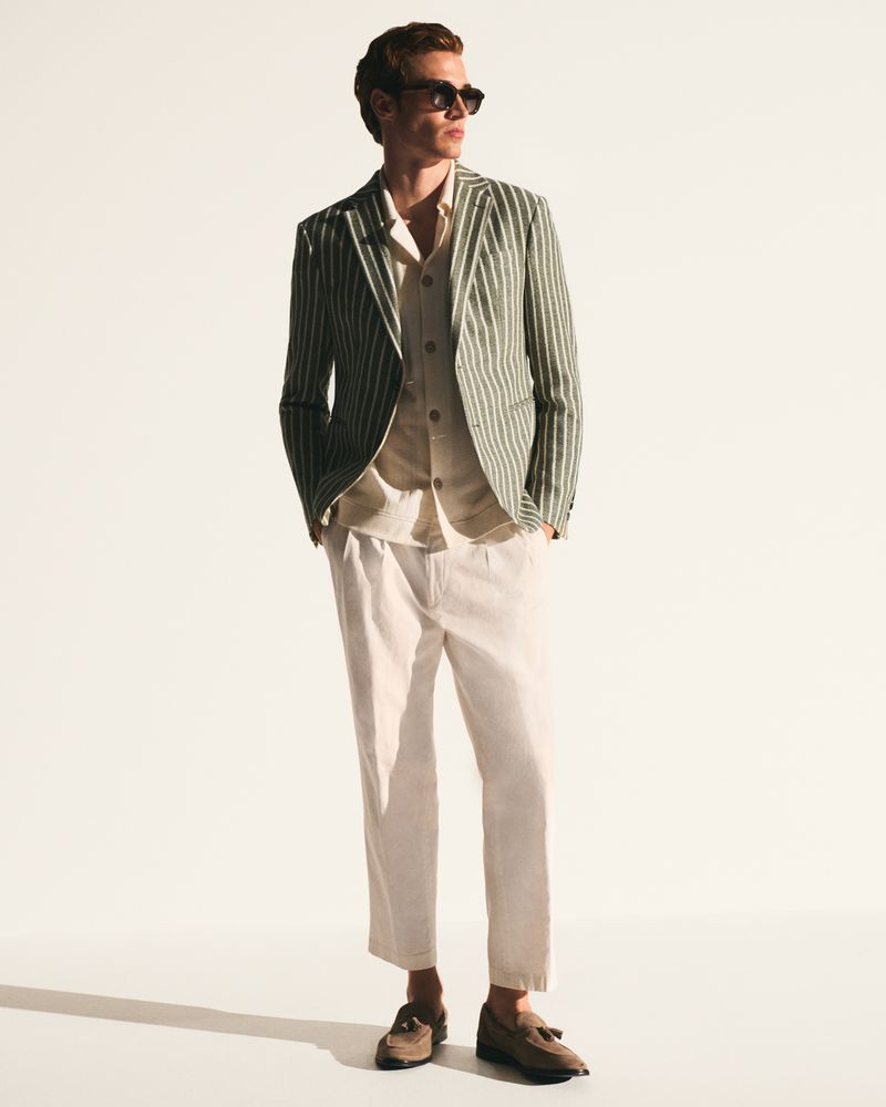 Man wearing a striped blazer, light shirt, white trousers, and sunglasses standing against a white studio background.