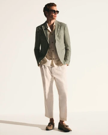 Man wearing a striped blazer, light shirt, white trousers, and sunglasses standing against a white studio background.