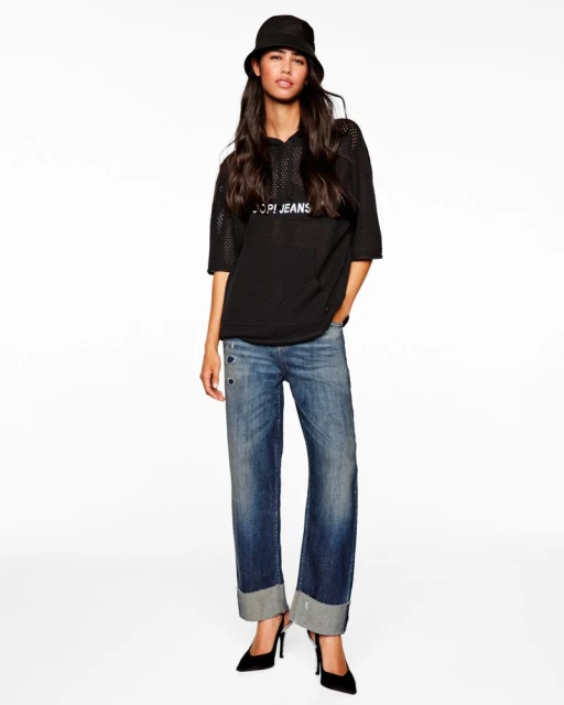 A woman stands in a black mesh hoodie, blue jeans, and black heels against a white backdrop, showcasing a casual fashion style.