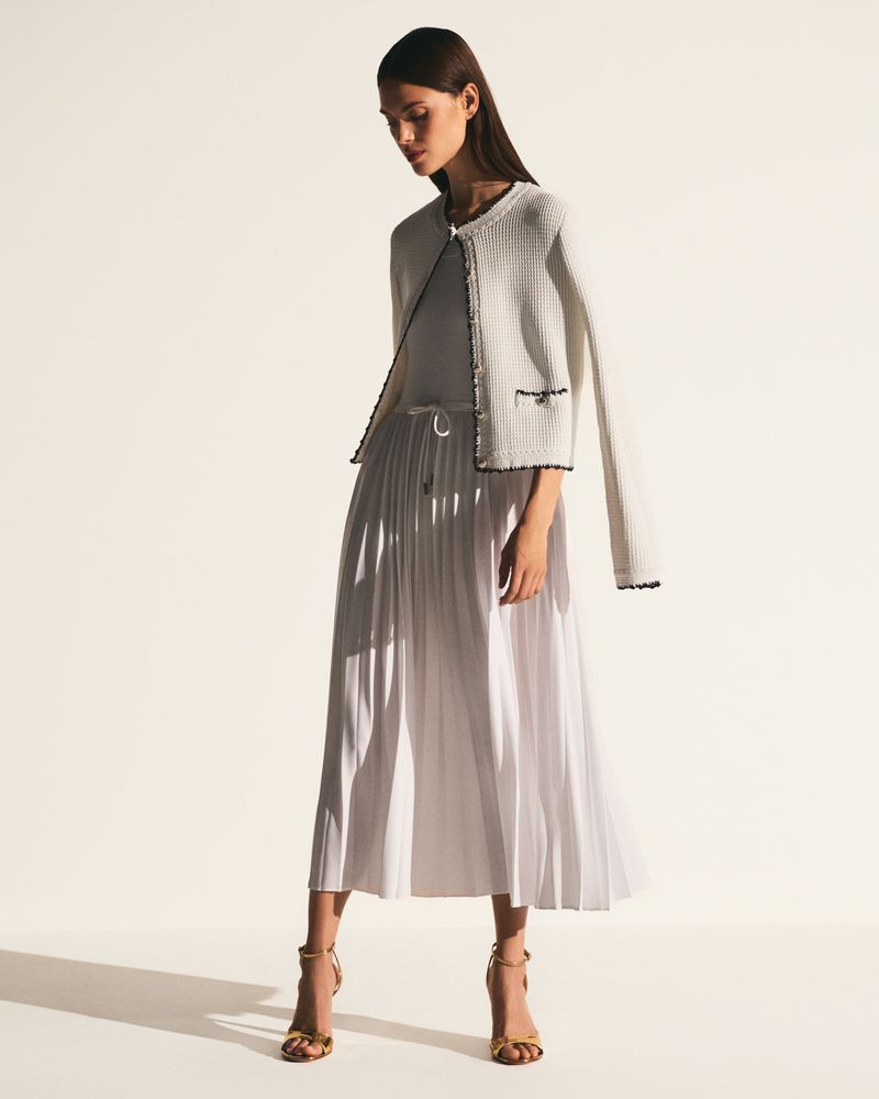 Full-length shot of a poised adult female model wearing a white jacket, a pleated midi skirt, and strappy heels against a bright backdrop.