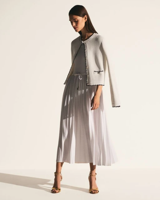 Full-length shot of a poised adult female model wearing a white jacket, a pleated midi skirt, and strappy heels against a bright backdrop.
