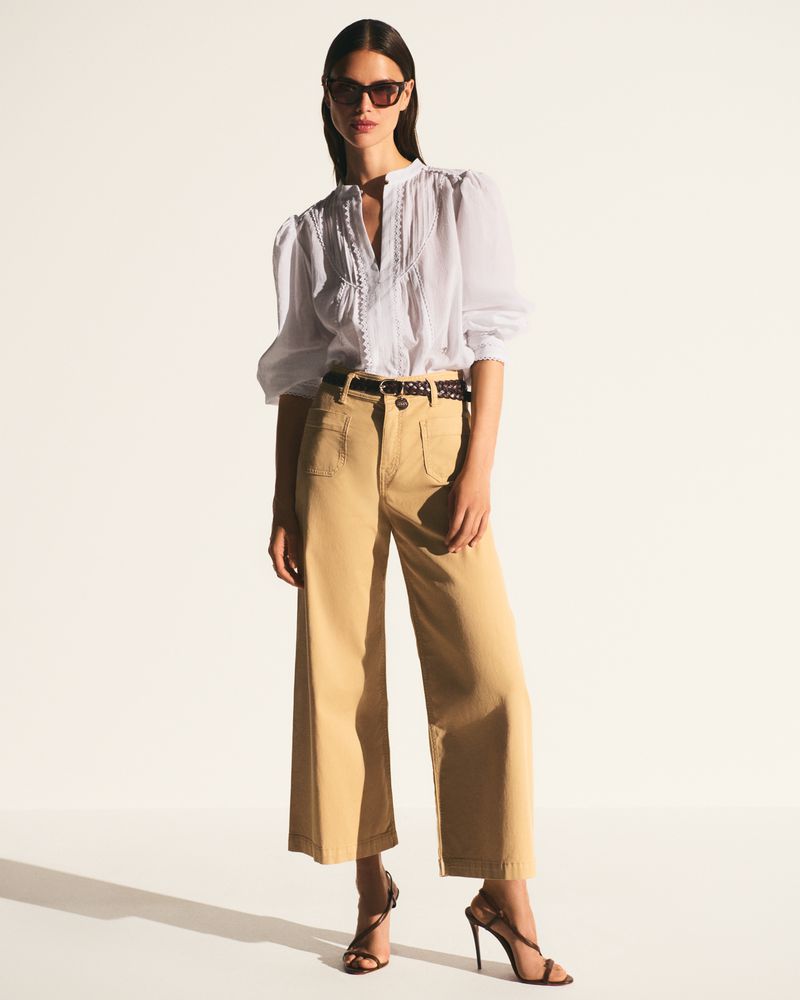 A stylish adult female poses in a white blouse, tan pants, and high heels against a bright white background in a fashion studio.