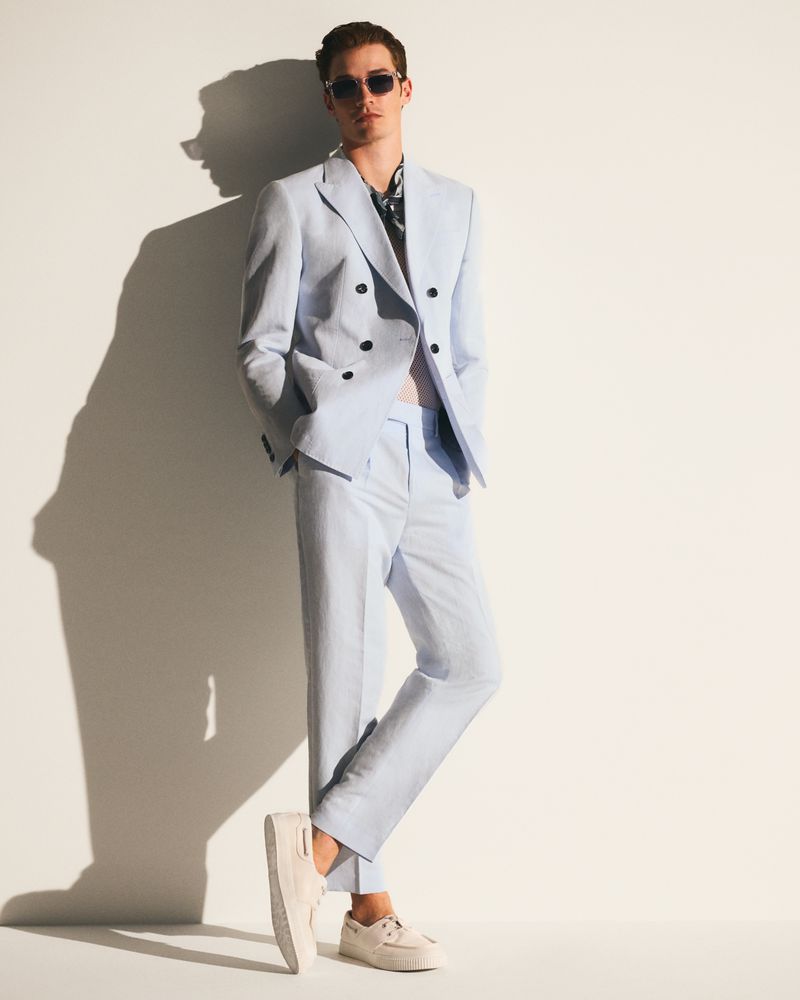 Man in a light blue suit and sunglasses poses against a white wall with a shadow. Fashion shot in an indoor setting.