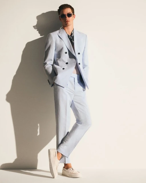Man in a light blue suit and sunglasses poses against a white wall with a shadow. Fashion shot in an indoor setting.