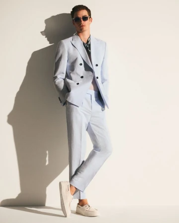 Man in a light blue suit and sunglasses poses against a white wall with a shadow. Fashion shot in an indoor setting.