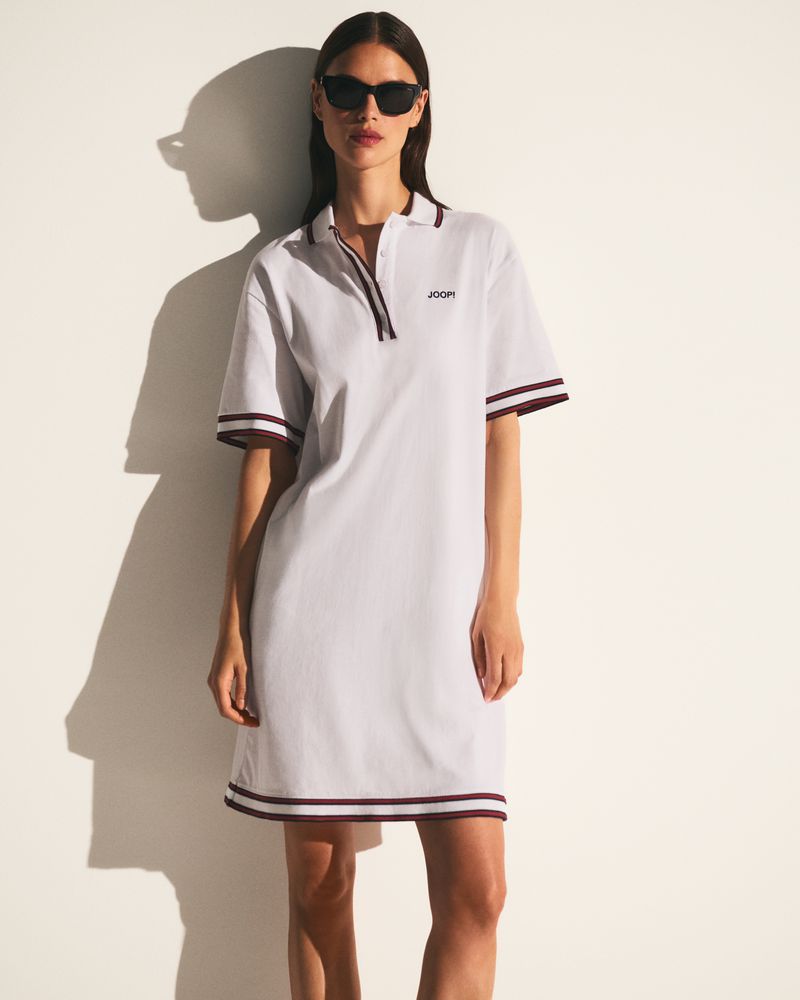 A woman poses in a white, short-sleeved polo dress with dark trim, accessorized with black sunglasses, against a bright white wall.