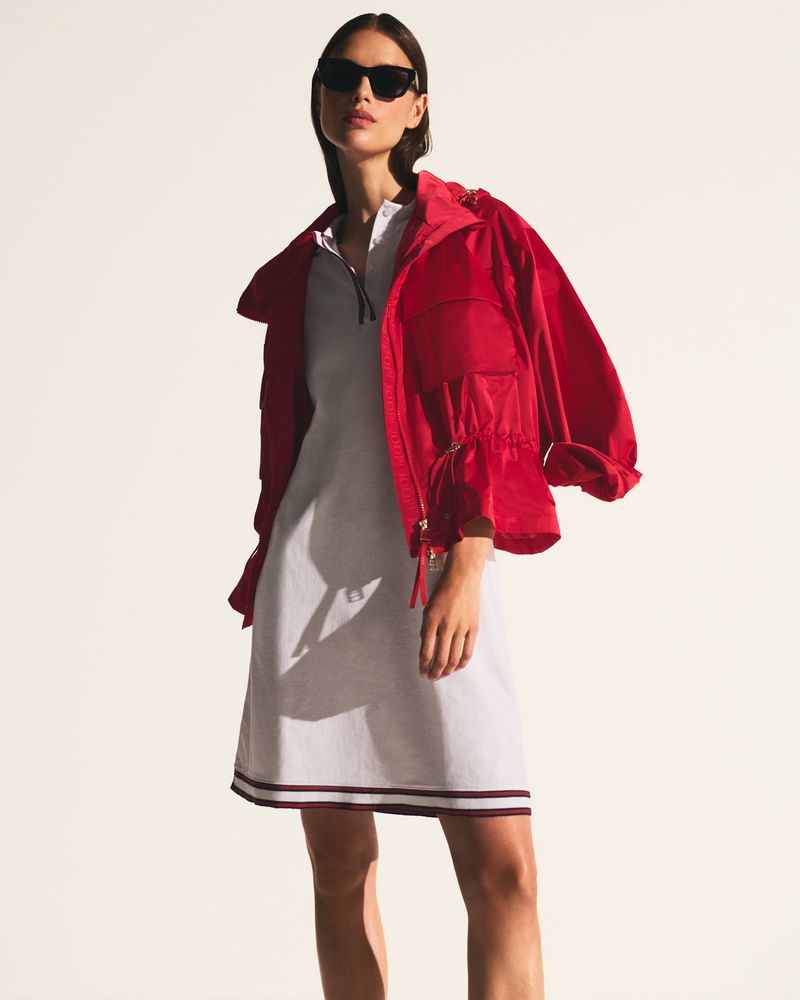 Model wearing dark sunglasses, a white polo dress, and an open red hooded jacket against a white studio background.