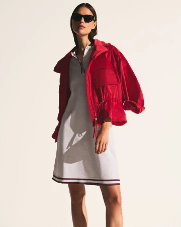 Model wearing dark sunglasses, a white polo dress, and an open red hooded jacket against a white studio background.