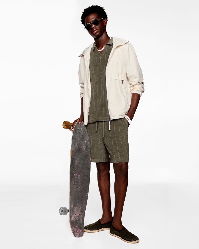 Young man in casual fashion poses with a skateboard against a white backdrop, wearing a jacket, matching shorts, polo shirt and sunglasses.