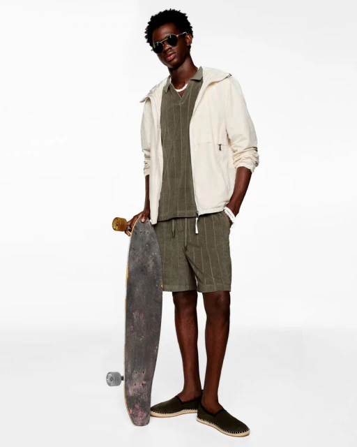 Young man in casual fashion poses with a skateboard against a white backdrop, wearing a jacket, matching shorts, polo shirt and sunglasses.