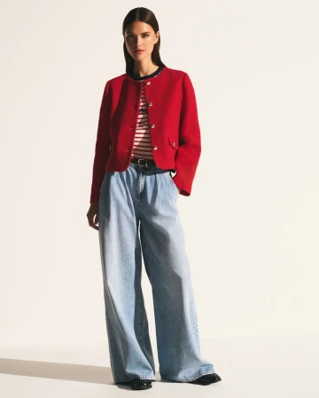 Model wearing a cropped red buttoned jacket, striped top, and wide-leg light blue jeans against an off-white seamless background.