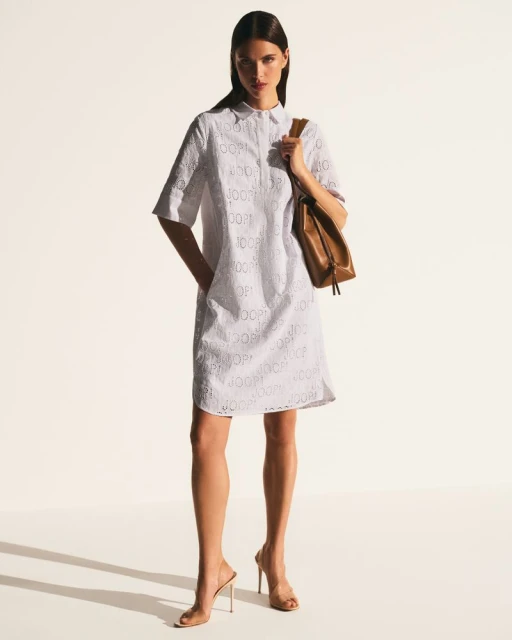 Fashion shot of a woman in a white eyelet dress with a brown leather purse on a white seamless backdrop, full length.