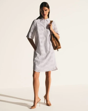 Fashion shot of a woman in a white eyelet dress with a brown leather purse on a white seamless backdrop, full length.