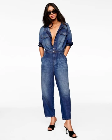 Model wearing a blue denim jumpsuit, black sunglasses, and black mule heels against a white seamless studio background.