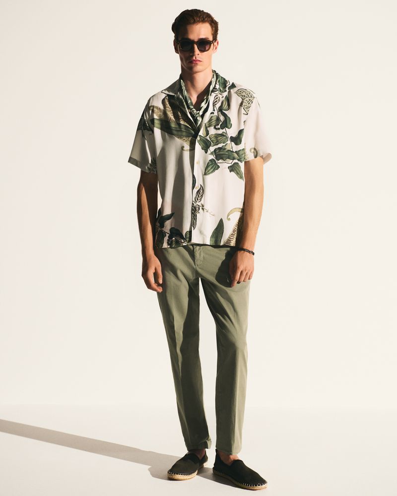 Full shot of an adult male wearing a short-sleeved shirt with a leafy pattern, olive pants, and sunglasses against a white backdrop.