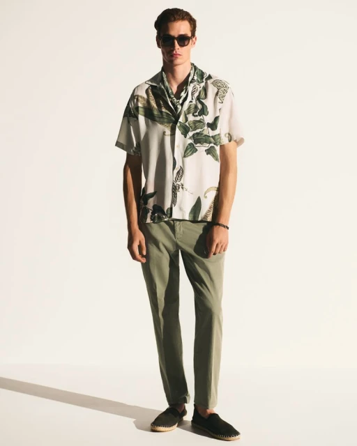 Full shot of an adult male wearing a short-sleeved shirt with a leafy pattern, olive pants, and sunglasses against a white backdrop.
