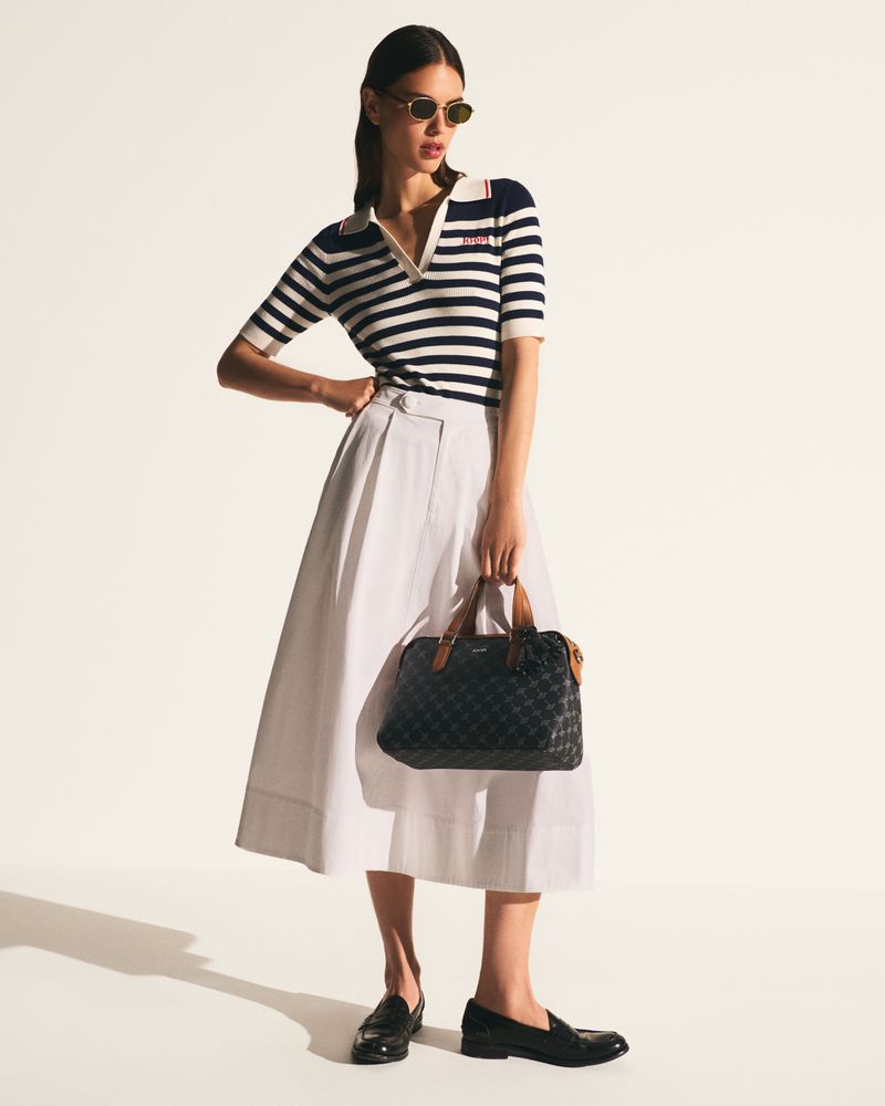 Model wearing a navy striped knit polo, white pleated midi skirt, sunglasses, and black loafers holding a patterned handbag in a studio.