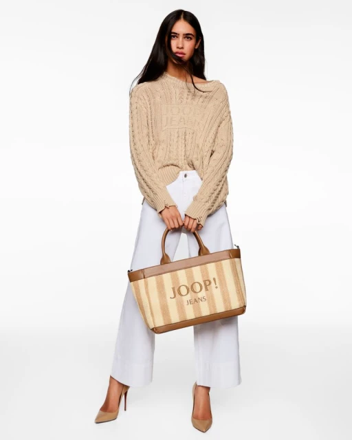 Brunette woman posing in a beige knit sweater, white wide-leg pants, and beige heels, holding a logo tote bag against white background.