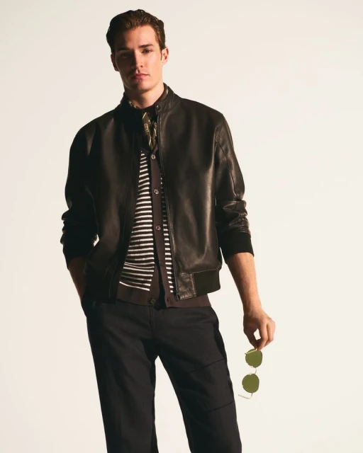 A fashionable adult male poses in a leather jacket, black pants, and striped sweater, holding sunglasses in a studio setting.