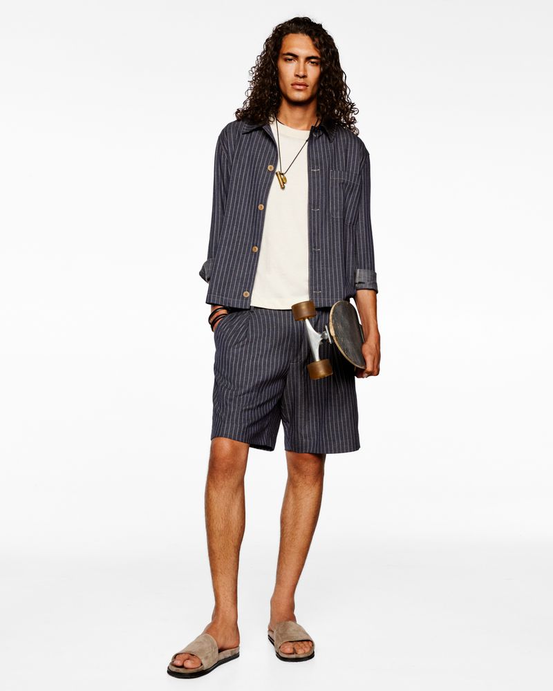 Young man in striped shirt and shorts holds a skateboard against a white background. Casual fashion, neutral colors, and long curly hair.