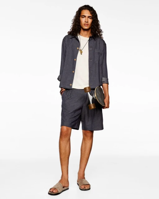 Young man in striped shirt and shorts holds a skateboard against a white background. Casual fashion, neutral colors, and long curly hair.