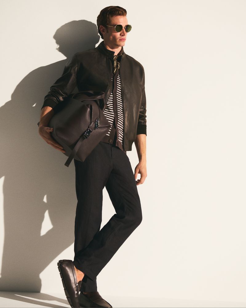 Man in a leather jacket carrying a brown duffel bag poses in a studio. He wears sunglasses, dark pants, and leather loafers.