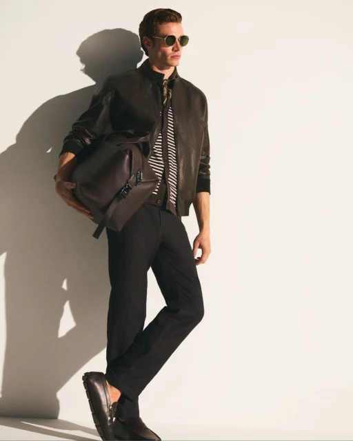 Man in a leather jacket carrying a brown duffel bag poses in a studio. He wears sunglasses, dark pants, and leather loafers.