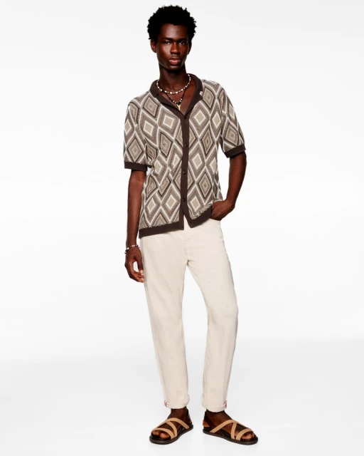 Full-length shot of a dark-skinned adult male posing in a patterned collared short-sleeved shirt, light jeans, and strappy sandals.