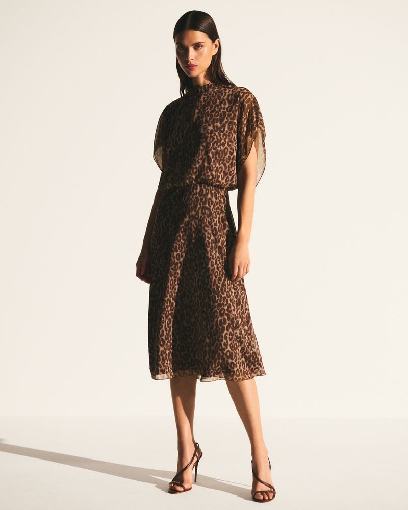 A brunette woman poses in a tan and brown animal print dress and strappy heels against a white background in a studio shot.
