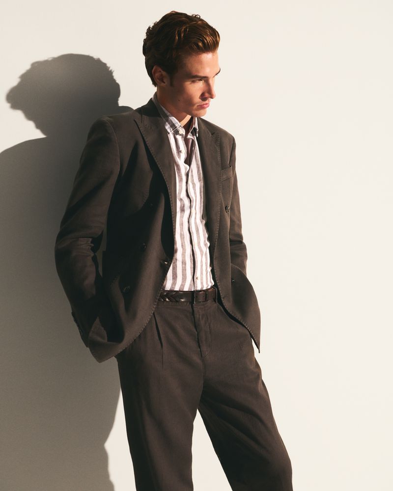 Man in a suit and striped shirt leans against a white wall. His hands are in his pockets and the lighting is soft and directional.