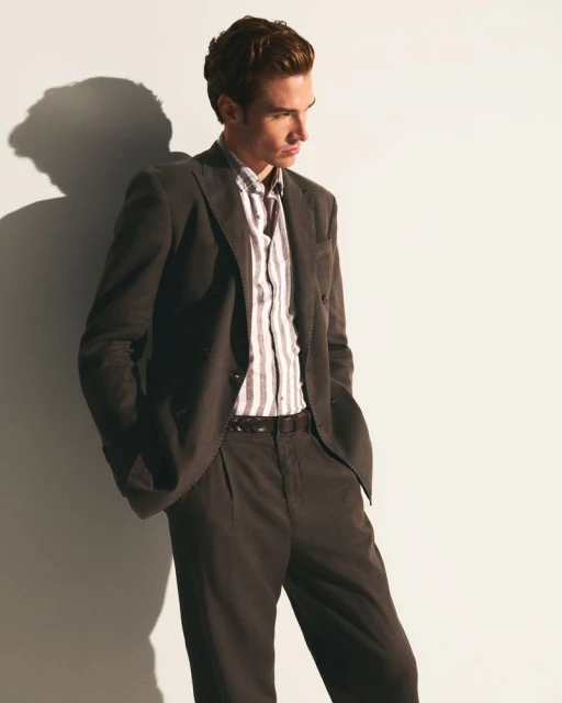 Man in a suit and striped shirt leans against a white wall. His hands are in his pockets and the lighting is soft and directional.