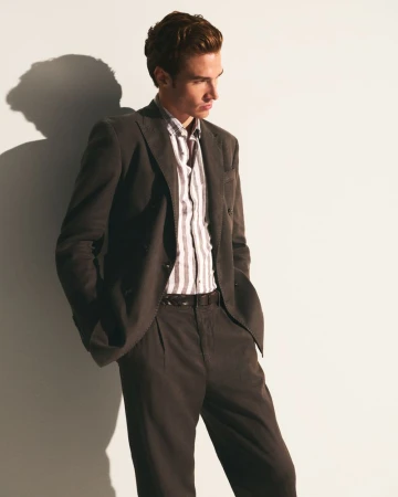 Man in a suit and striped shirt leans against a white wall. His hands are in his pockets and the lighting is soft and directional.