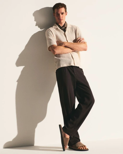 Full shot of a young adult male leaning against a white wall in a casual shirt, dark pants, and sandals in a brightly lit indoor setting.