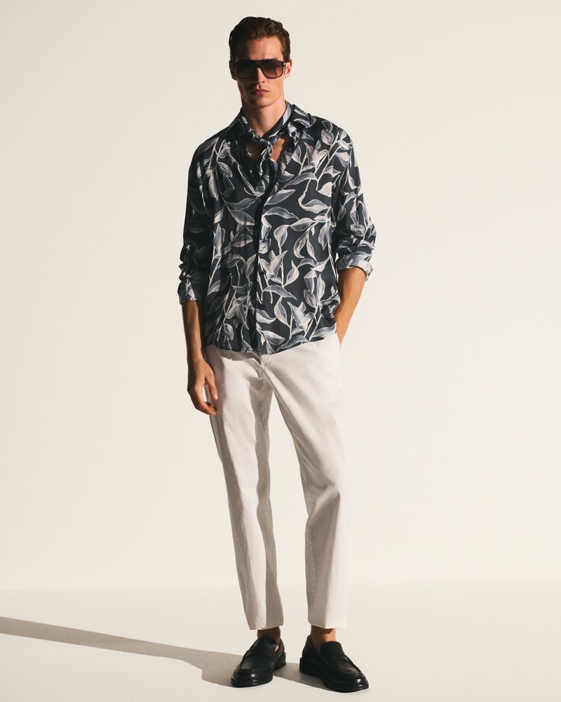 Man in a patterned shirt and light pants stands in a photo studio wearing sunglasses and black loafers looking at the camera.