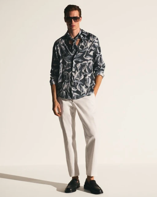 Man in a patterned shirt and light pants stands in a photo studio wearing sunglasses and black loafers looking at the camera.