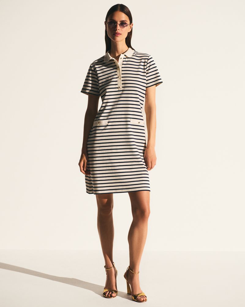 Model wearing a navy and white striped polo dress with gold high heel sandals against a clean white backdrop.