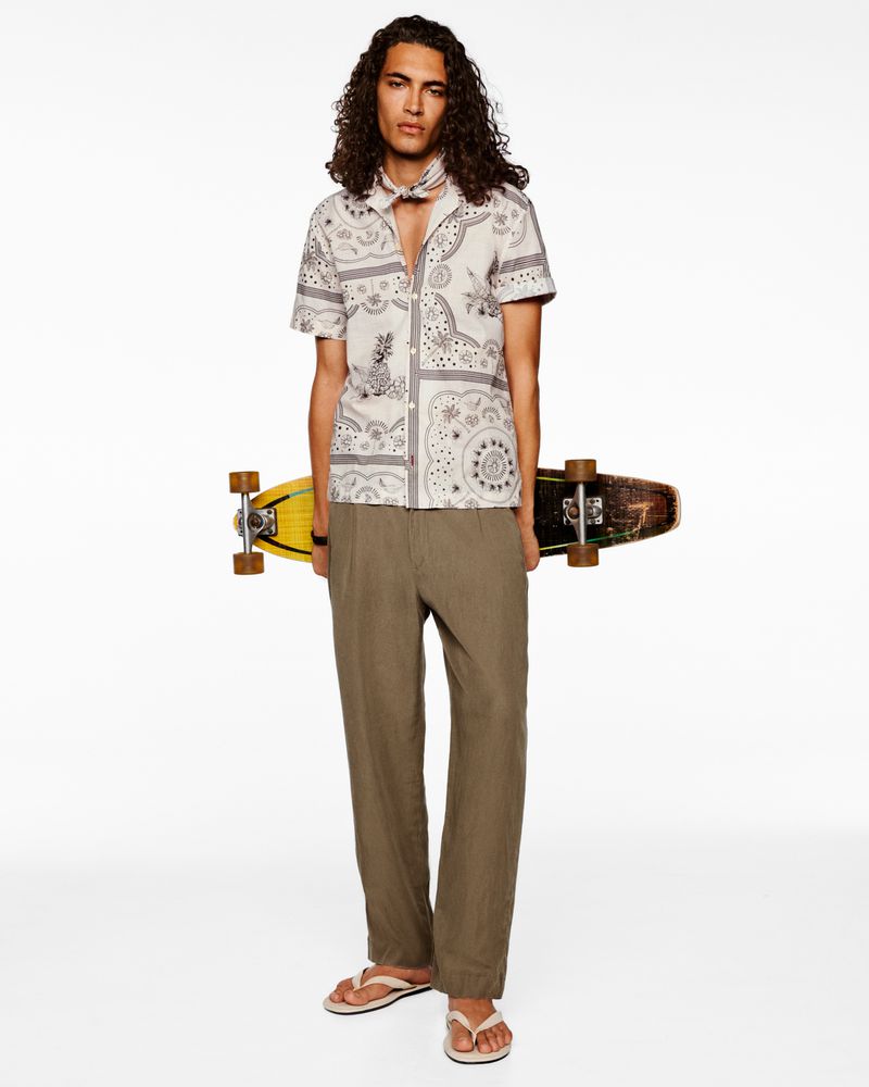 A slender adult male with long curly hair stands in a photo studio holding a skateboard while wearing a patterned shirt and earth-tone pants.