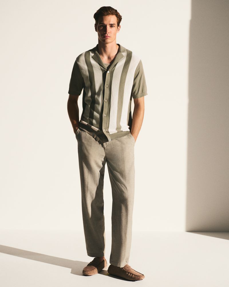 A full shot of a fair-skinned male model wearing a striped collared shirt, light-colored pants, and moccasins against a plain white backdrop.