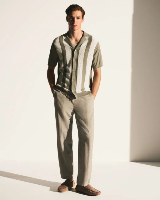 A full shot of a fair-skinned male model wearing a striped collared shirt, light-colored pants, and moccasins against a plain white backdrop.