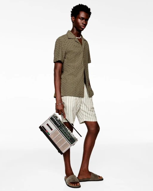 Full-length shot of a dark-skinned male model in an olive-green collared shirt, striped shorts, and slides holding a boombox against white.