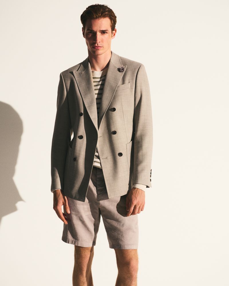 A young adult male model poses indoors, wearing a tan double-breasted blazer, striped shirt, and light tan shorts in a fashion shoot.