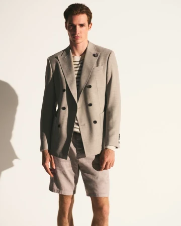 A young adult male model poses indoors, wearing a tan double-breasted blazer, striped shirt, and light tan shorts in a fashion shoot.