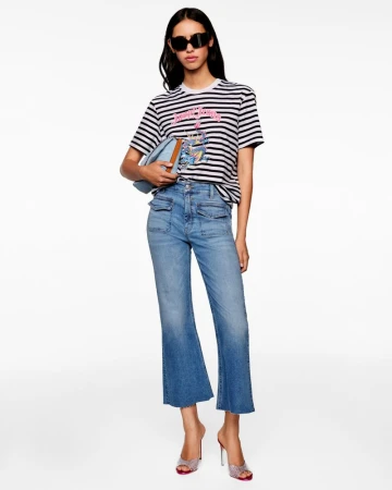 Model wearing a striped t-shirt, cropped flared blue jeans, dark sunglasses, and metallic heeled mules against a white background.