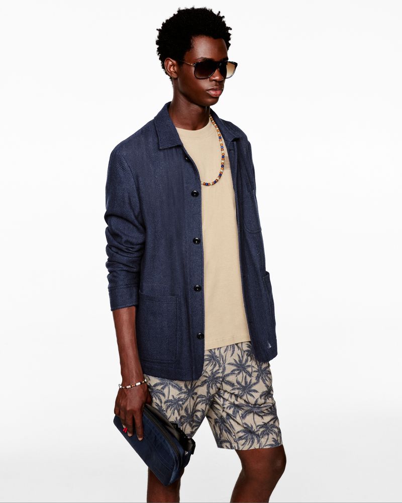 A young Black man poses in a casual outfit, featuring a beige T-shirt, patterned shorts, a navy jacket, sunglasses, and a beaded necklace.